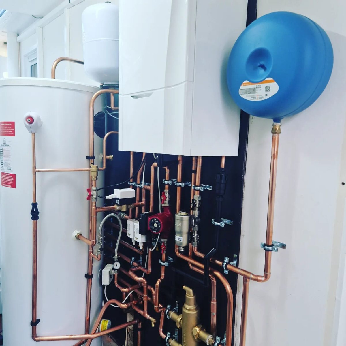 Wall-hung boiler beside an unvented cylinder with a pump, zone valves and an expansion vessel on a black backboard